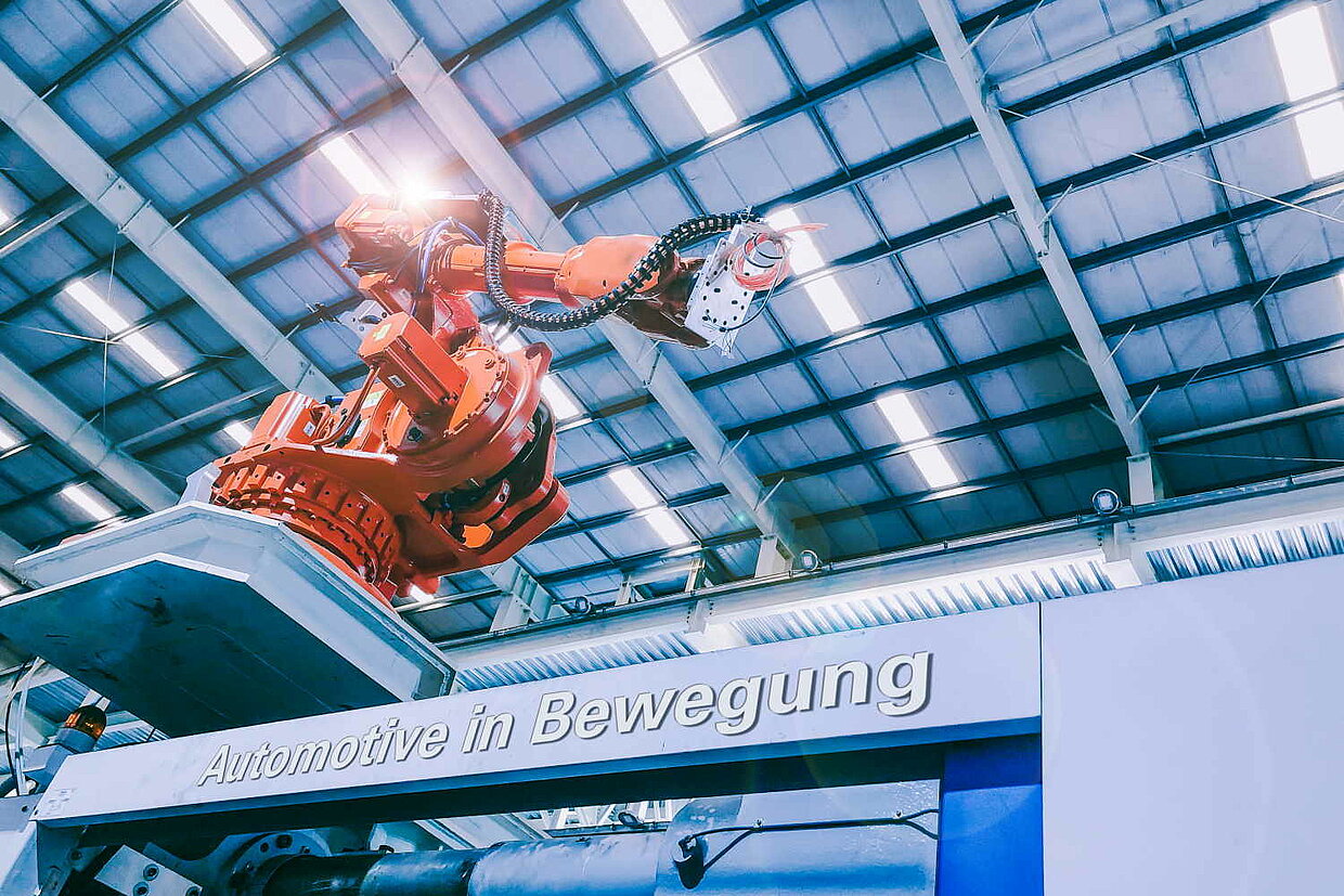 Veranstaltung "Automotive in Bewegung" Roboterarm sitzt erhöht in einer Produktionshalle. Auf der Anlage steht "Automotive in Bewegung" geschrieben.